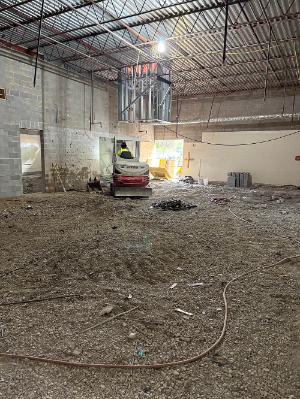 Interior renovation site with exposed roof trusses, unfinished masonry walls, loose gravel, debris, and hoses across the floor.