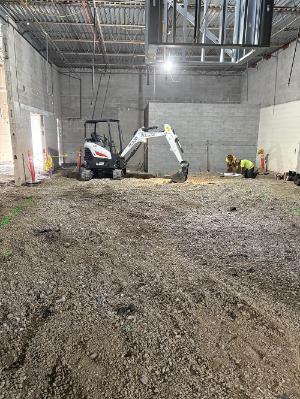 Interior construction site with a compact excavator on a gravel floor and two workers in high-visibility clothing working beside a partially excavated area. Exposed masonry walls and steel roof framing surround the space.