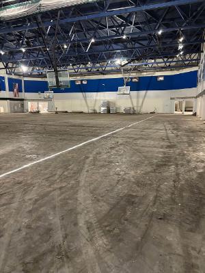 Large indoor gymnasium under renovation, with a rough unfinished floor marked by construction equipment tracks, multiple basketball hoops, blue and white wall bands, and exposed roof trusses.
