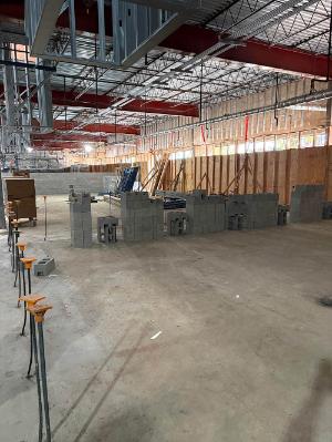 Interior of a building under construction, with exposed steel roof framing, HVAC ductwork, unfinished walls, and stacks of concrete masonry blocks on the floor.