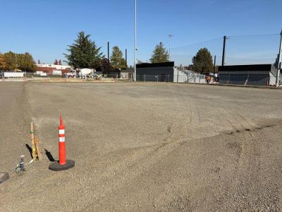 Outdoor construction area with a broad gravel surface, orange traffic delineator, utility stakes, and equipment in the background.
