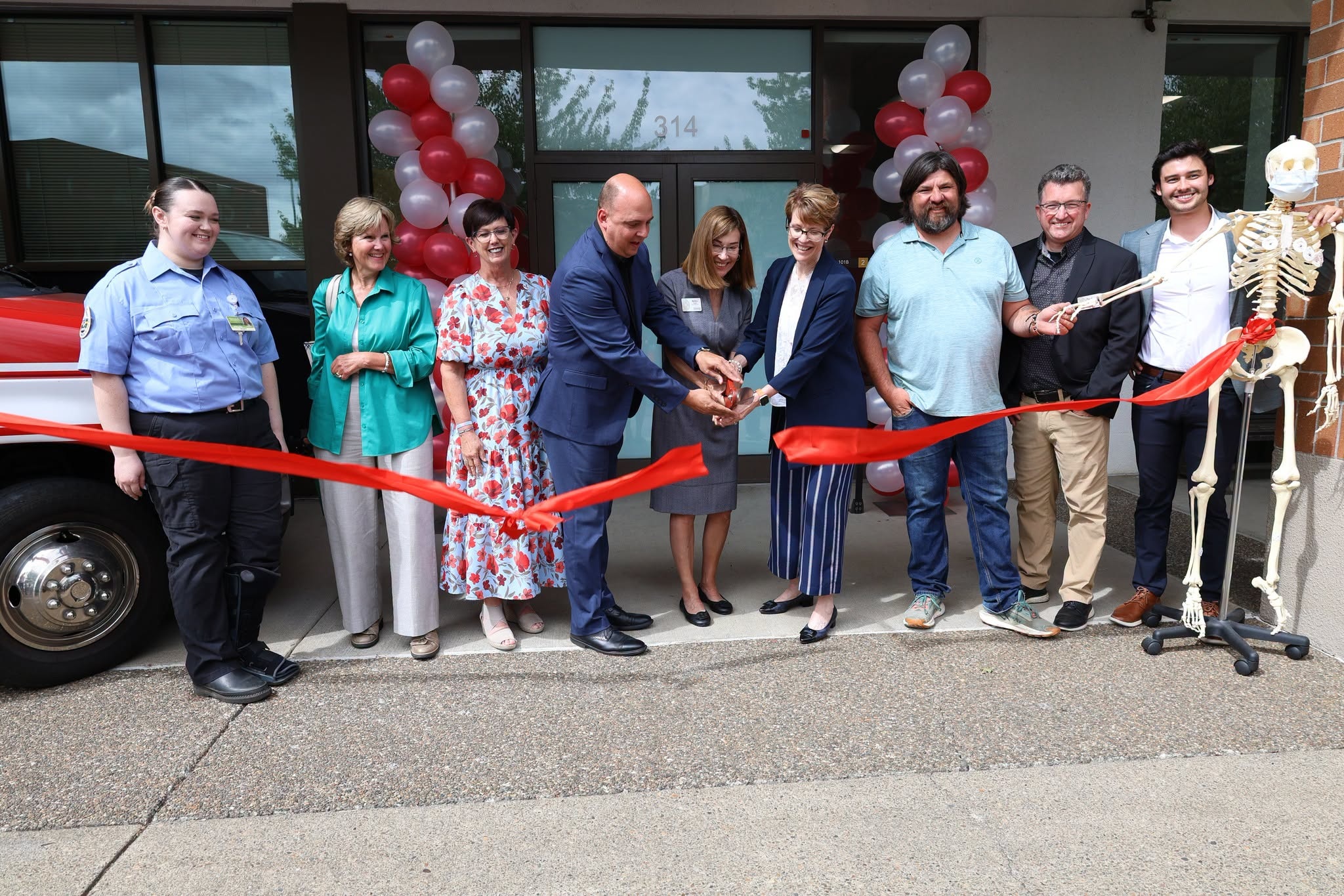 A group of people cutting a ceremonial grand opening red ribbon with giant scissors in front of a college building