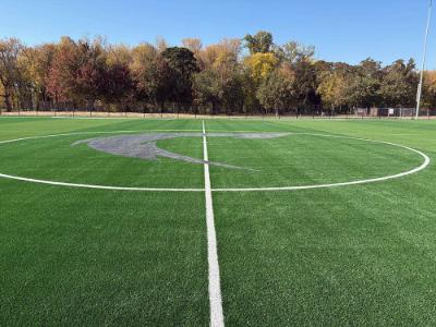 Completed artificial turf soccer field with white centerline markings and a large gray stylized letter 
