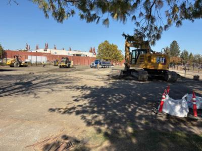 Outdoor construction site with a yellow excavator beneath a tree, several pieces of heavy equipment, and a dump truck on graded ground beside a brick building.