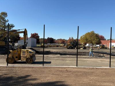 Outdoor sports court construction site with a yellow road roller in the foreground, additional heavy equipment and workers in the background, and vertical steel posts along the graded ground.