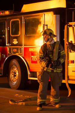 A firefighting student with an uncharged hose on his shoulder looks in the direction of a fire at nighttime. A fire engine is in the background illuminated by the flames.