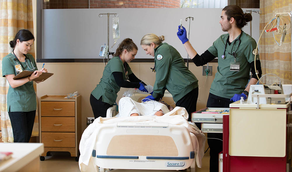 Four Chemeketa nursing students training in a simulation lab. Two of the students work on a medical dummy in a hospital bed, another is making notes on a chart, and another is preparing a syrringe.